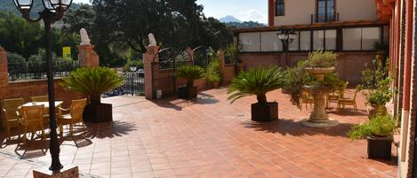 Un patio con suelo de baldosas, mobiliario exterior, plantas en macetas y un edificio con tejado de tejas.