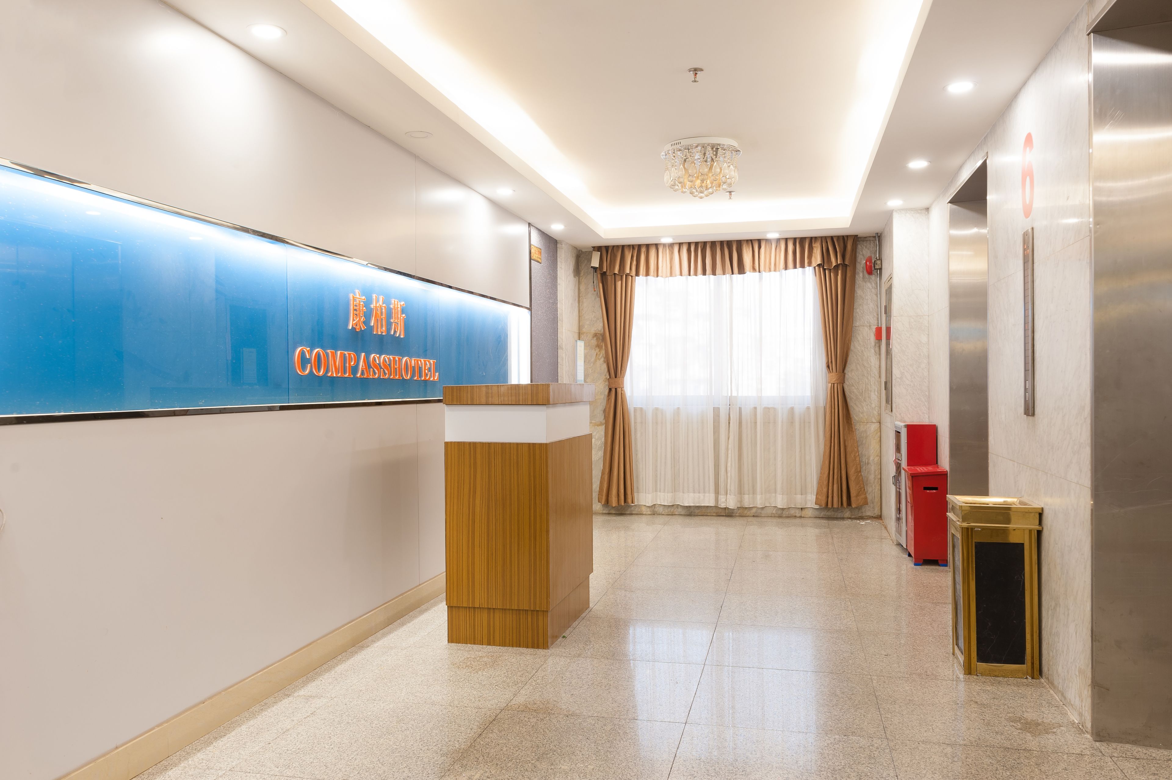 A hotel reception area with a blue sign, a wooden reception desk, a chandelier, and a red trash bin.