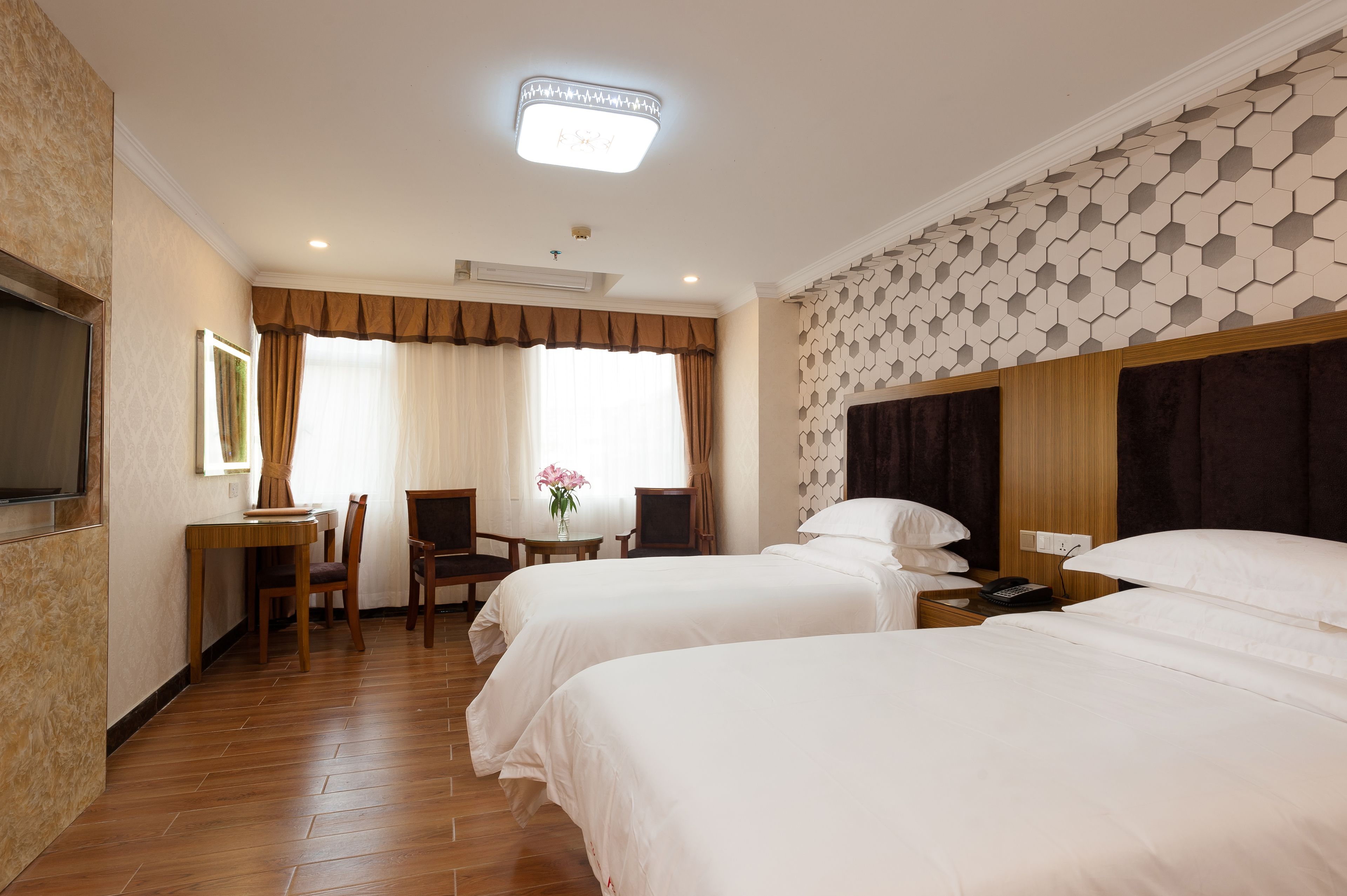 A hotel room with two beds, a desk with a chair, a small table with a vase, and a patterned wall.