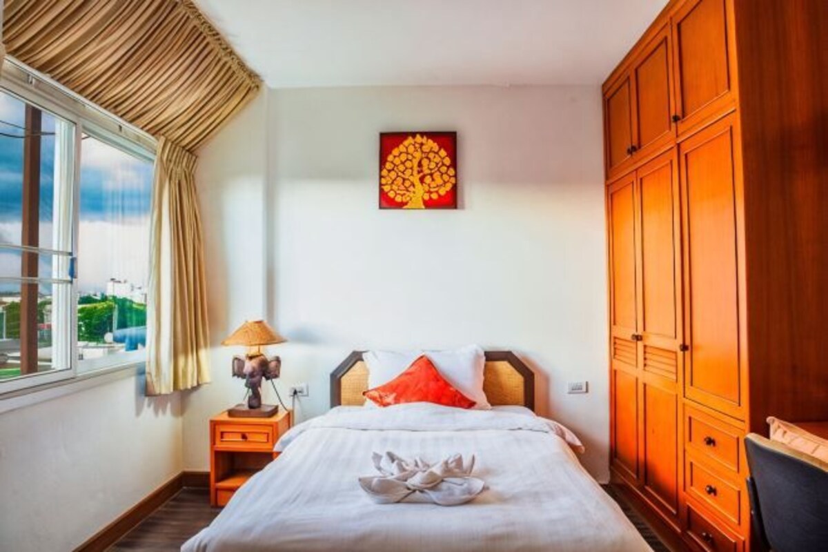 A bedroom with a bed, a nightstand, a lamp, a window with curtains, a wooden wardrobe, and a wall art piece.