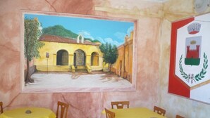 A dining area with yellow tables and chairs, a mural of a yellow building, and a coat of arms on the wall. - Rosa Hotel (Capoterra)