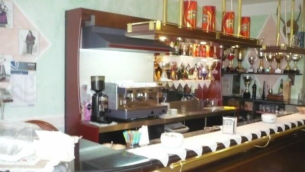 A bar area with a coffee machine, shelves with bottles, and a counter with a glass display. - Rosa Hotel (Capoterra)