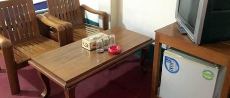Una mesa de madera con una caja y un objeto rojo, dos sillas de madera, un televisor sobre un soporte, y un refrigerador con calcomanías.