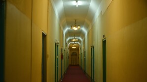 Un largo y estrecho pasillo con alfombra roja, bordeado por puertas a ambos lados, que conduce a un par de puertas dobles al final.
