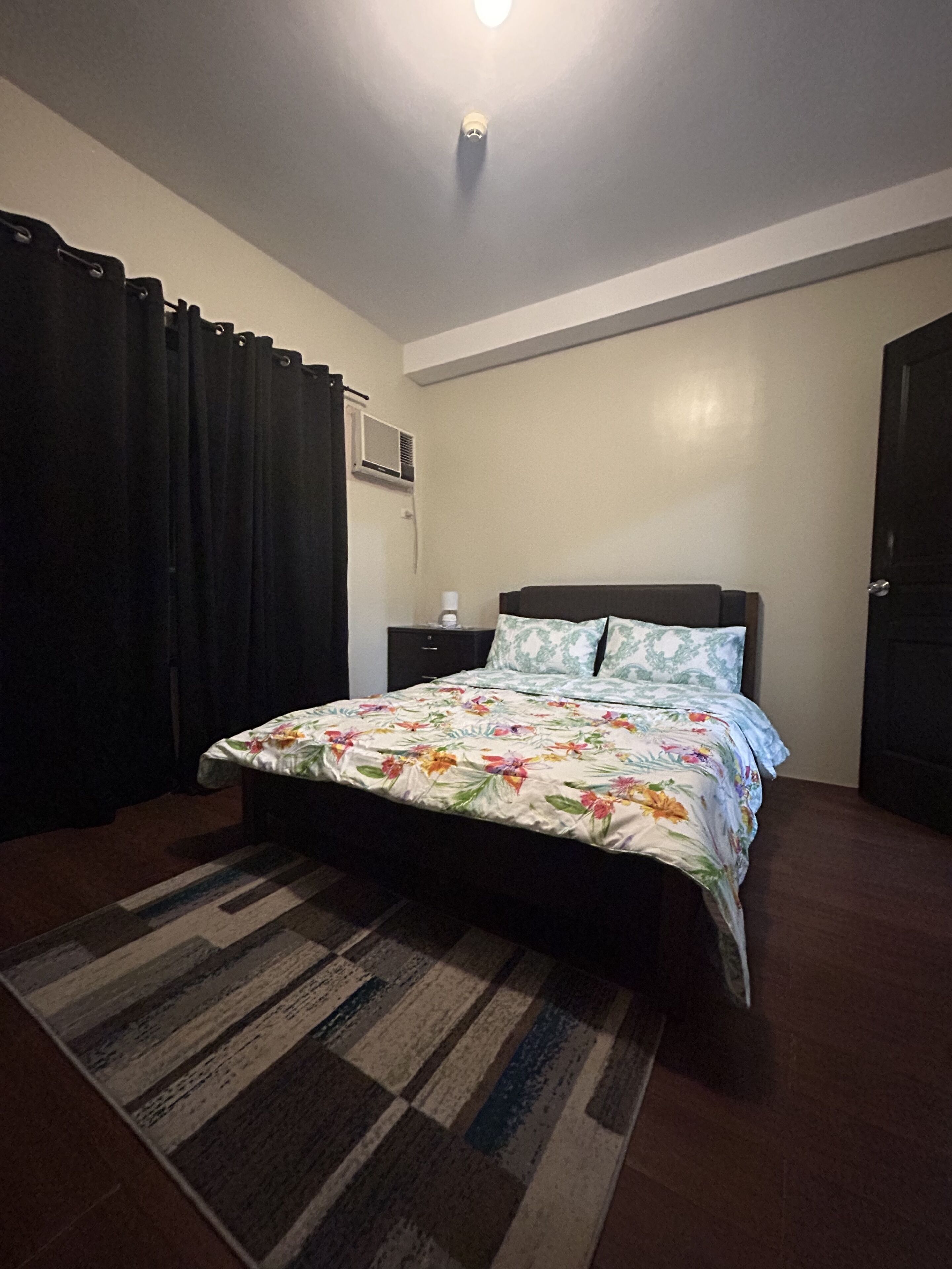 Un dormitorio con una cama, una mesita de noche, aire acondicionado de pared y una puerta.