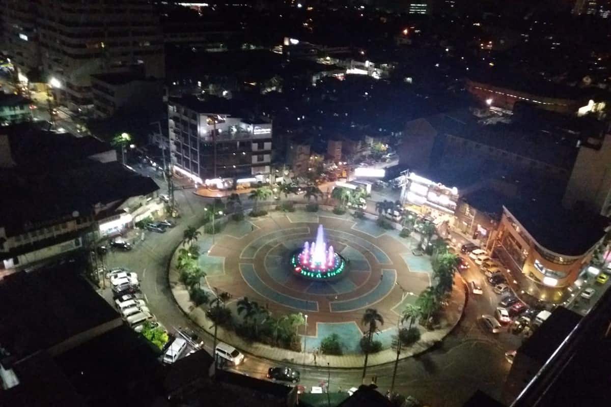 Vista nocturna de una plaza urbana con una fuente central, rodeada de edificios y coches estacionados.
