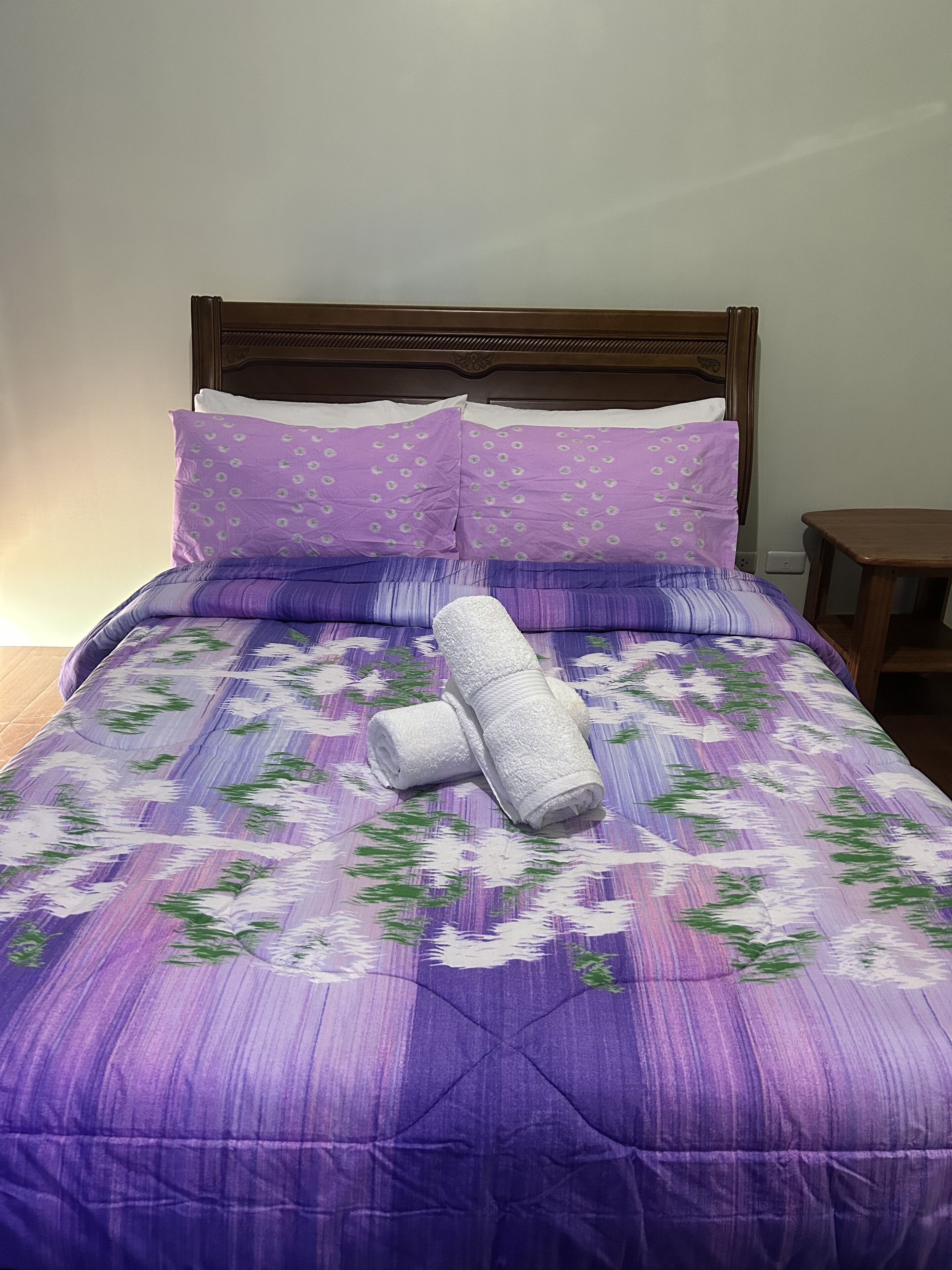 Una cama con una colcha morada estampada con flores, dos almohadas con un diseño parecido, y dos toallas blancas enrolladas.