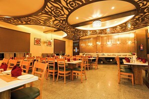 A restaurant with multiple tables and chairs, a decorative ceiling, and a kitchen area in the background. - Hotel Silk City (Varanasi)