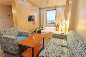 A hotel room with a sofa, two armchairs, a small wooden table, a TV mounted on the wall, and a window with a city view. - Hotel Silk City (Varanasi)
