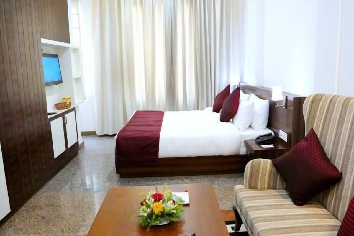 Ein Hotelzimmer mit einem Bett, einer Couch, einem Couchtisch mit Blumen, einem Fernseher und Vorhängen.