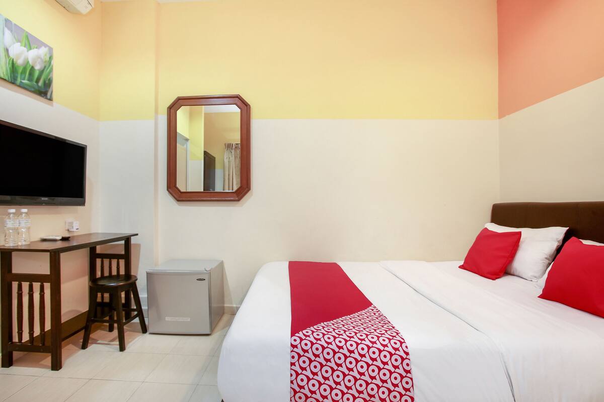 A hotel room with a bed, a desk, a chair, a small refrigerator, a TV, and a mirror.