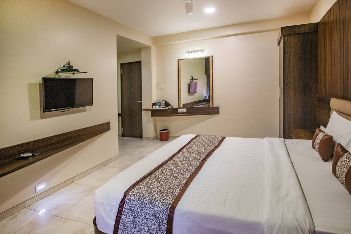 A hotel room with a large bed, a flat-screen TV mounted on the wall, a vanity with a mirror, and a wooden headboard.