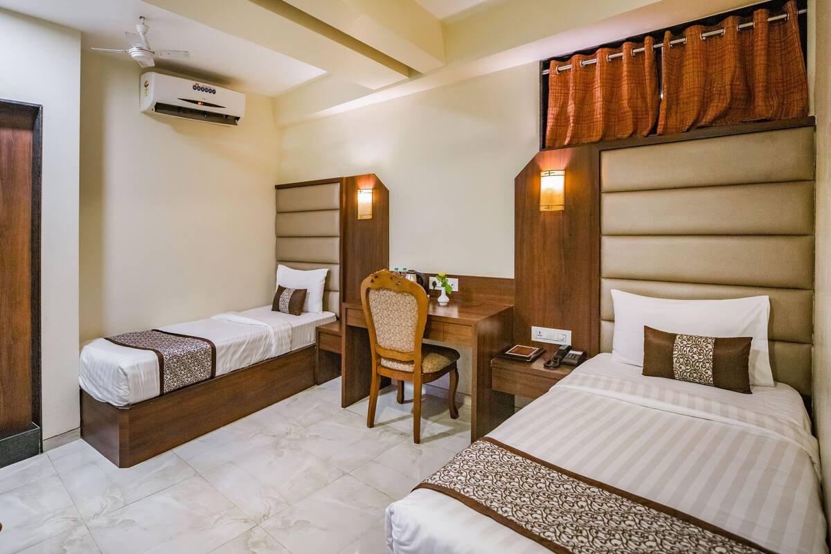 A hotel room with two single beds, a desk with a chair, an air conditioning unit, and a wooden wardrobe.