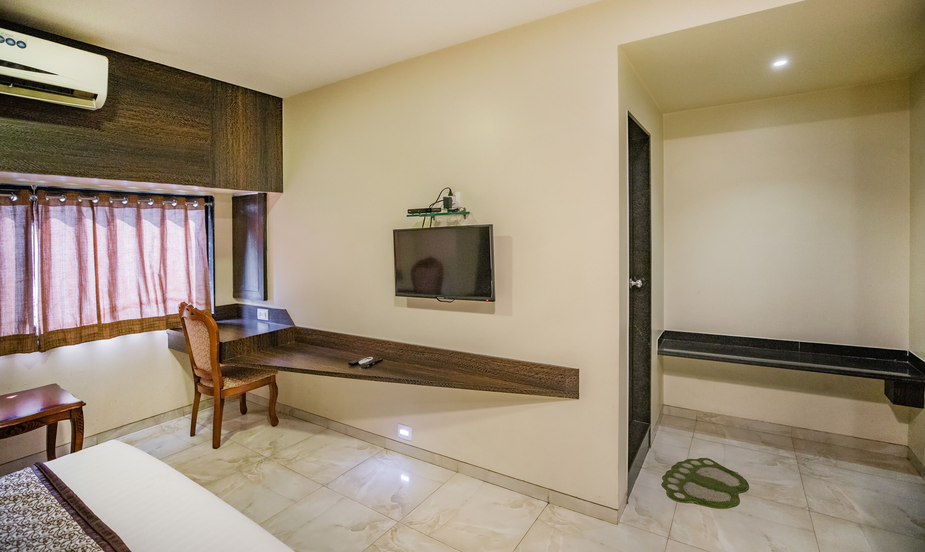 Habitación de hotel con una cama, una silla de madera, un escritorio con computadora, un televisor montado en la pared y un baño con ducha e inodoro.