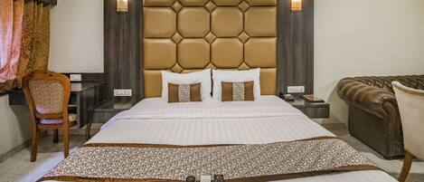 A hotel room with a large bed, a headboard with a geometric pattern, two wall-mounted lamps, a desk with a telephone, and a brown leather armchair.