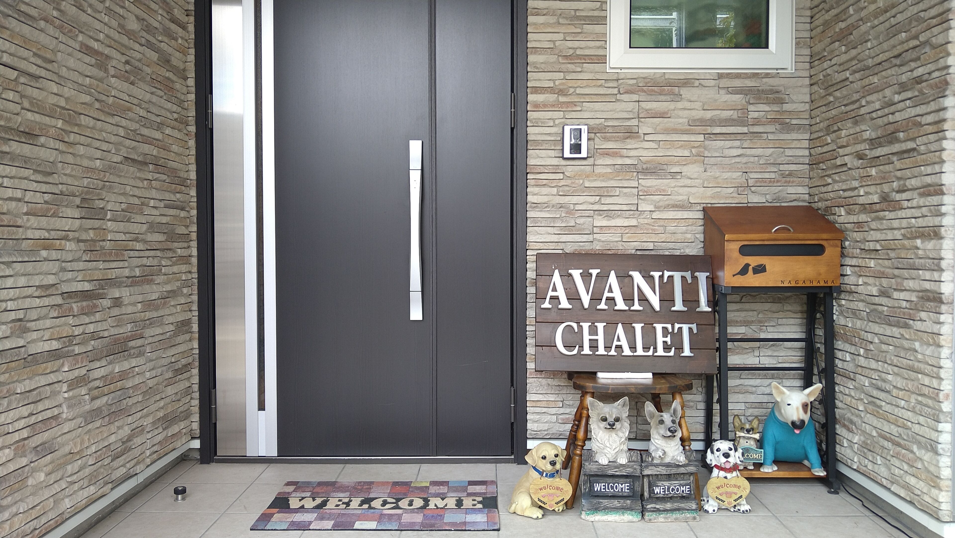 A modern building entrance with a dark door, a welcome mat, a sign reading "AVANTI CHALET", and decorative dog sculptures.