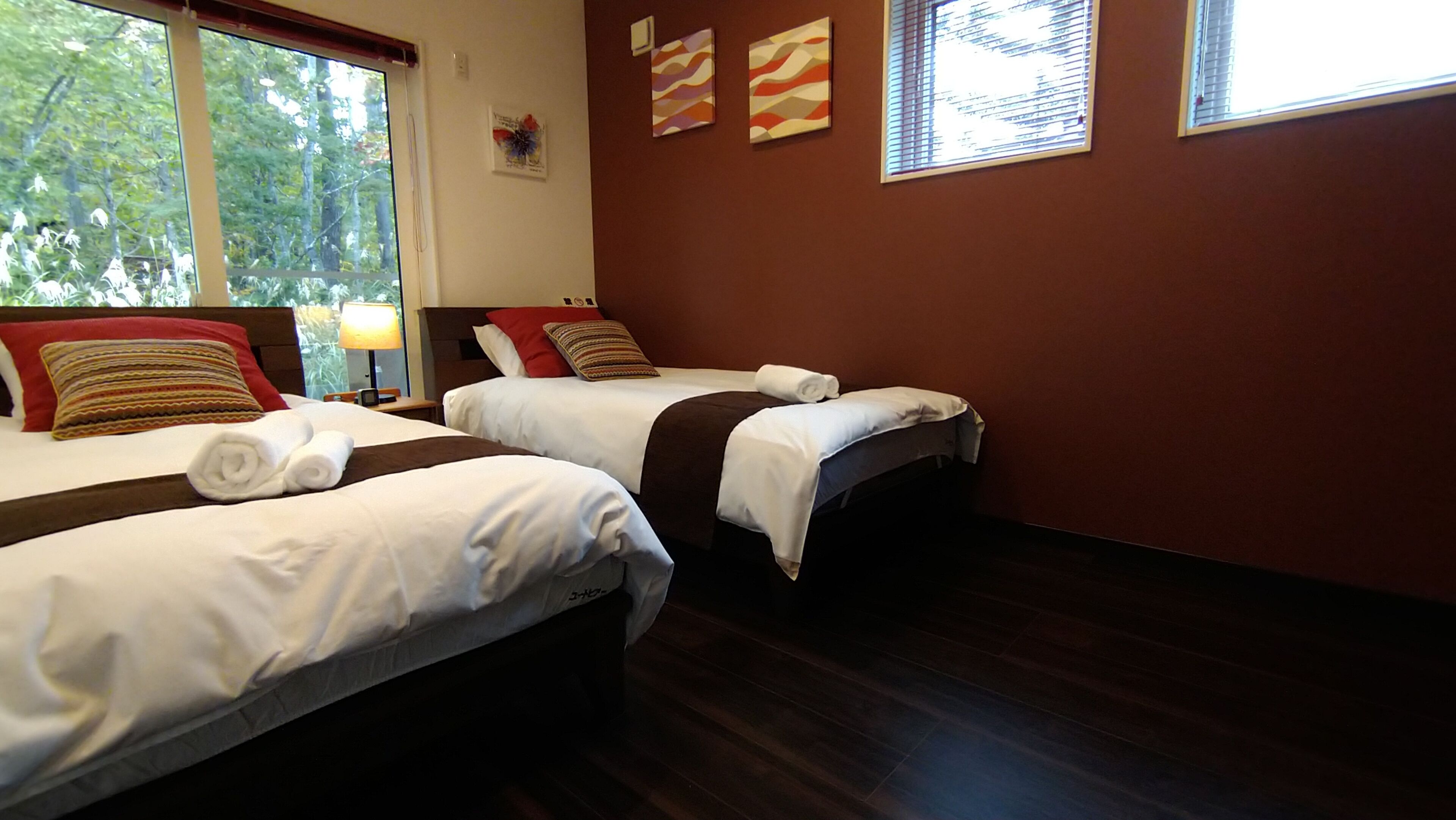 A hotel room with two beds, each with white linens and brown headboards. There are two windows with blinds, and a lamp on the left side of the room.