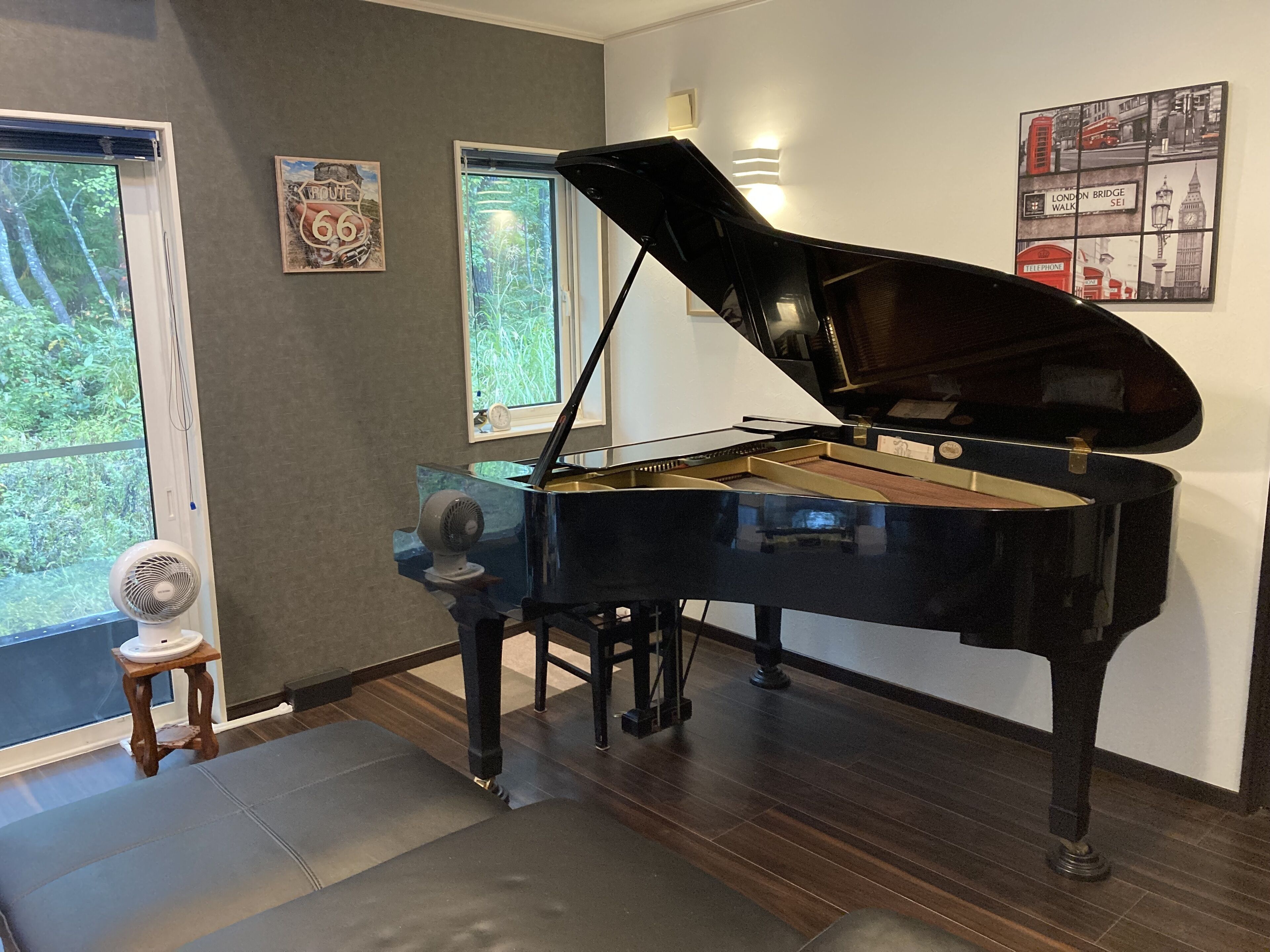 Un piano de cola en una habitación con un ventanal grande, un pequeño ventilador y un cuadro en la pared.