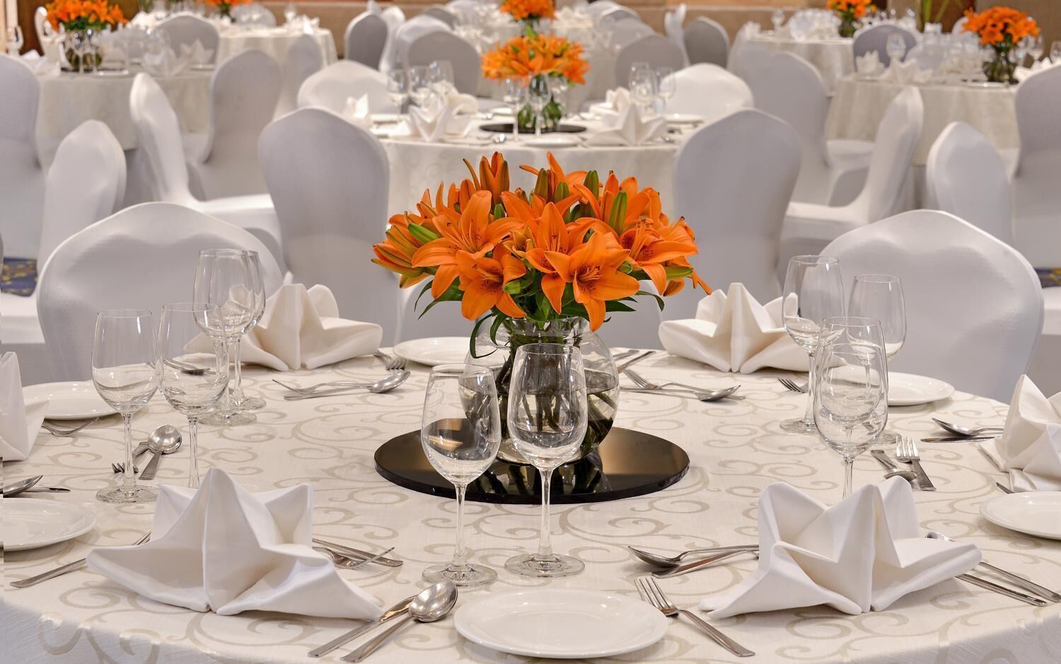 A banquet table set with white tablecloths, white chairs, and a centerpiece of orange flowers in a clear glass vase.