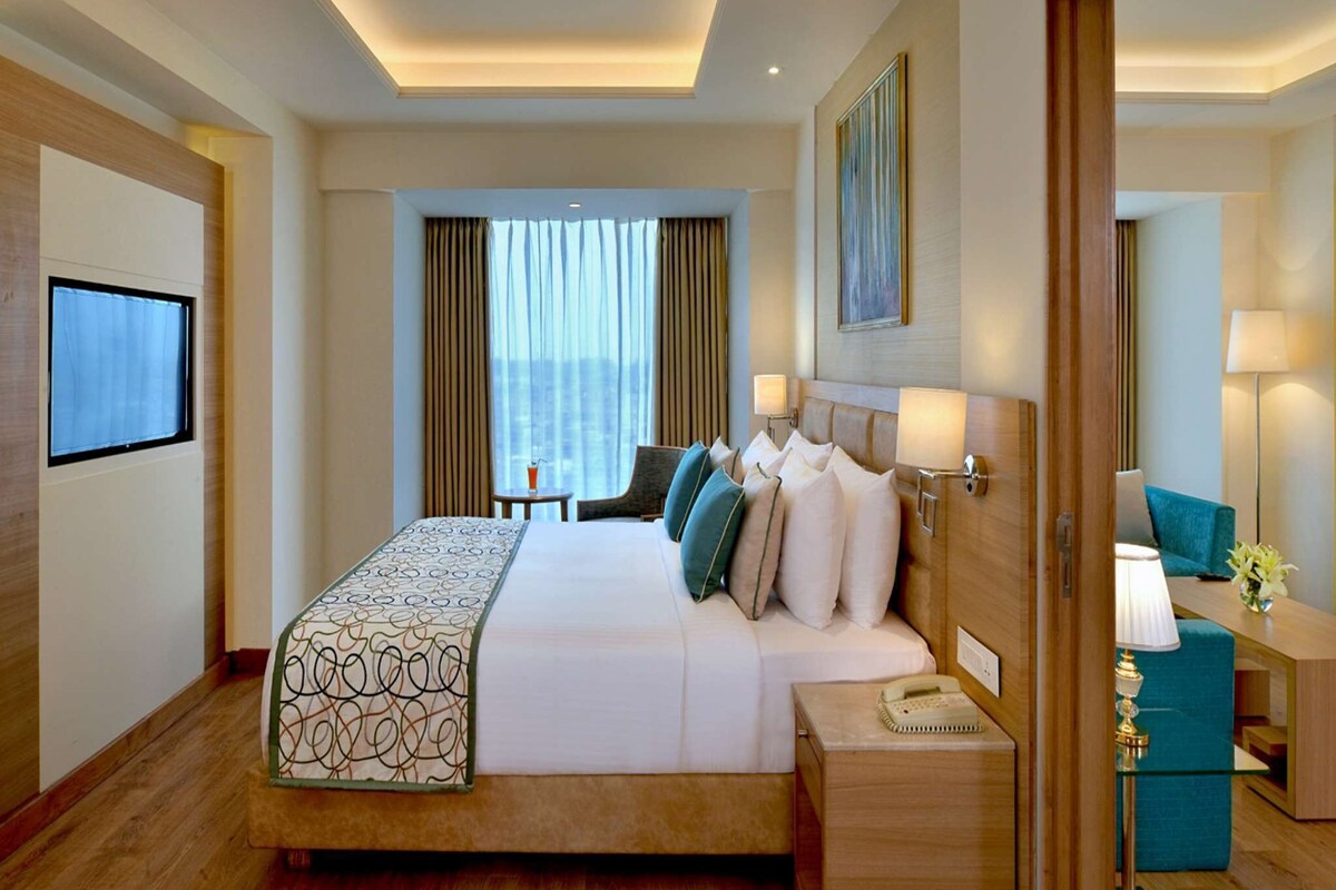 A hotel room with a large bed, bedside lamps, a desk with a lamp, and a view of the city through the window.