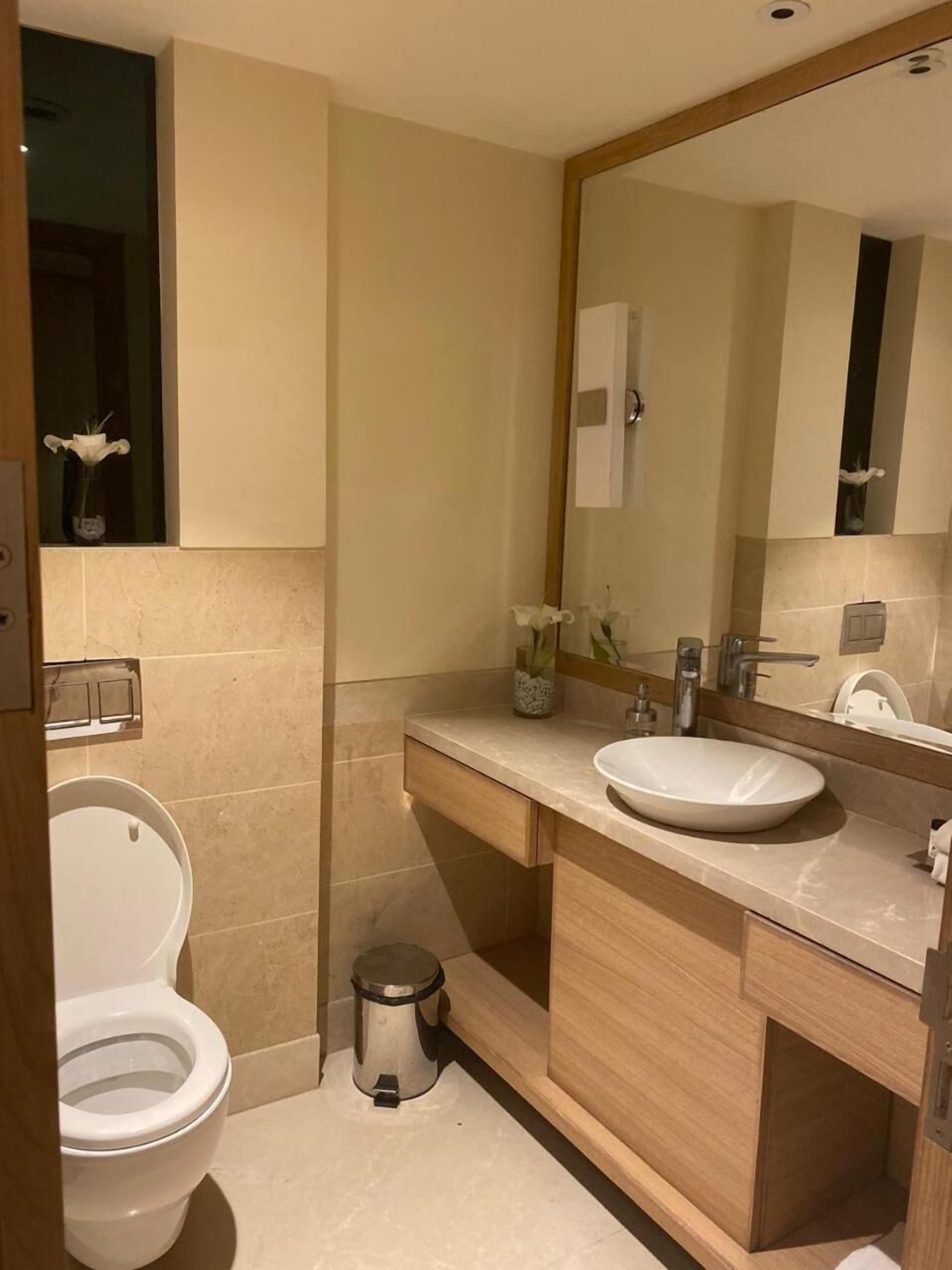 Un baño moderno con inodoro blanco, lavamanos con un espejo grande y un bote de basura pequeño.