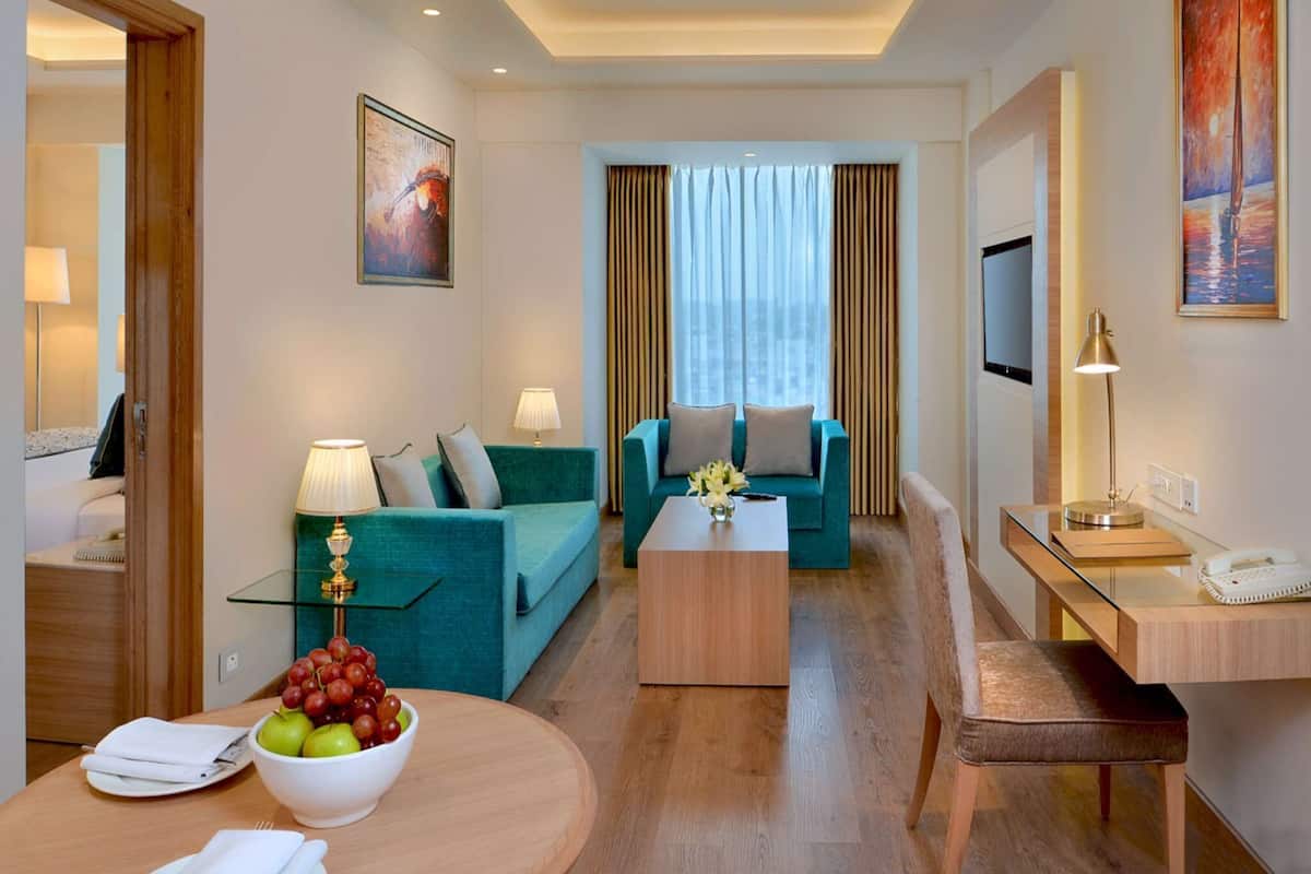 A modern hotel room with a teal sofa, a wooden coffee table, a glass side table, a desk with a lamp, a TV, and a painting on the wall.