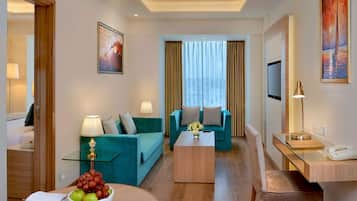 A modern hotel room with a teal sofa, a wooden coffee table, a glass side table, a desk with a lamp, a TV, and a painting on the wall.