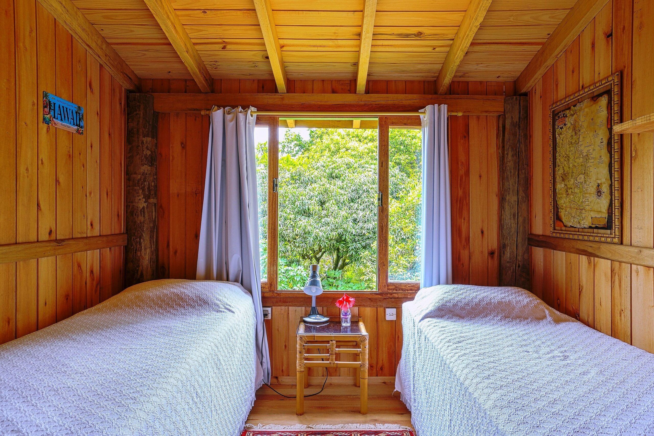 Una habitación tipo cabaña de madera con dos camas, una mesa con una lámpara y un ventanal con cortinas.