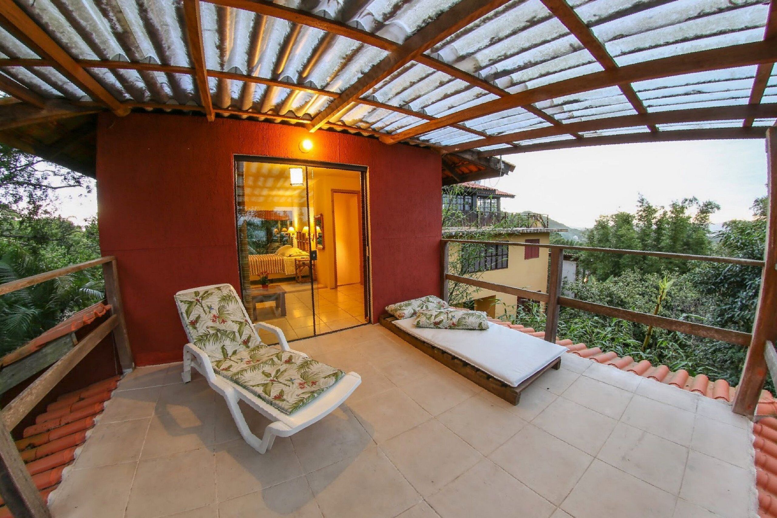 A covered balcony with a red wall, a white chair, a small table, and a bed with pillows.