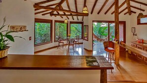 A spacious room with a high ceiling and exposed wooden beams, large windows offering views of lush greenery, a dining area with a wooden table and chairs, and a hammock on a balcony. - Studios do Barão (Imbituba)