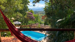 A hammock overlooking a swimming pool with a view of a lush landscape and distant mountains. - Studios do Barão (Imbituba)