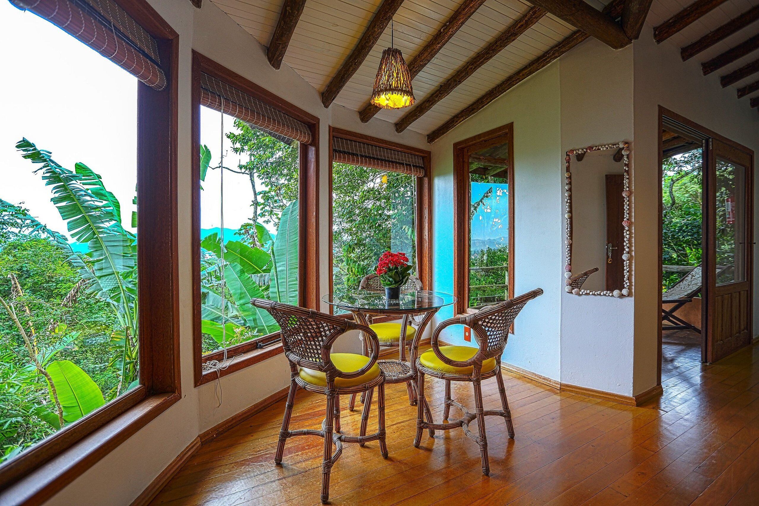 Una habitación con ventanas amplias, sillas de mimbre, una mesa redonda y una lámpara colgante.