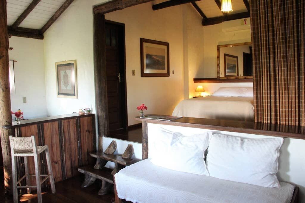Ein gemütliches Zimmer mit einer hölzernen Bank, einem Bett mit weißen Leinen und einem Spiegel, der das Bett und ein Bild an der Wand widerspiegelt.