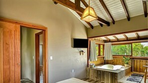 A bedroom with a large bed, wooden beams, a ceiling fan, a TV, a window with curtains, and a small bathroom area with a bathtub. - Studios do Barão (Imbituba)
