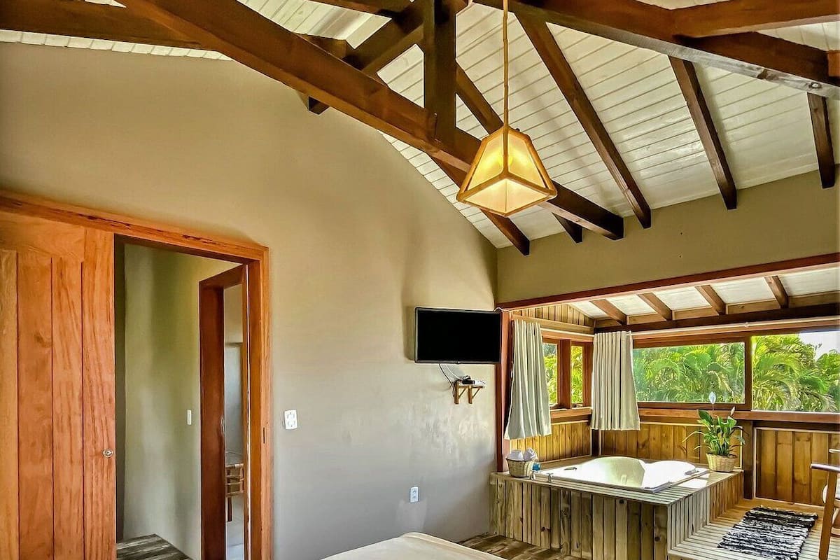 A bedroom with a large bed, wooden beams, a ceiling fan, a TV, a window with curtains, and a small bathroom area with a bathtub.