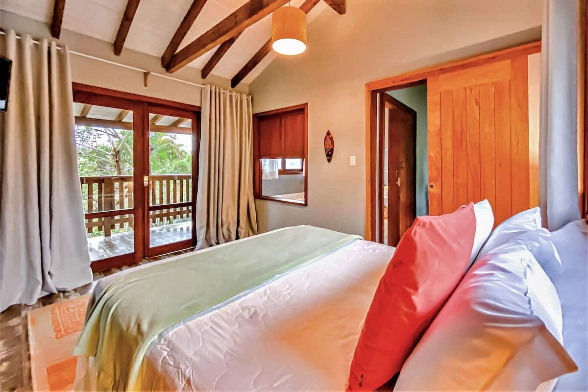 A bedroom with a bed, wooden beams, a window with curtains, and a door.