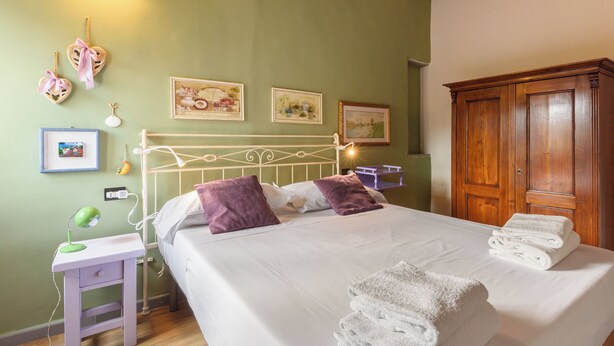 A neatly made bed with white linens and purple pillows, a bedside table with a lamp, and a wooden wardrobe. - Una Finestra sulle Cappelle Medicee by Mmega (Florence)