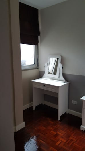 A small, white desk with a mirror and a window with a dark blind. The room has a wooden floor and light gray walls.