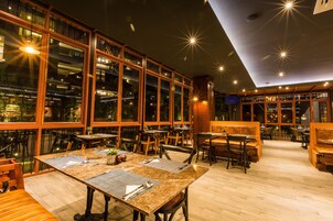 Un restaurante con techos altos, amplias ventanas y mobiliario de madera. Las mesas están dispuestas con manteles individuales y cubiertos.