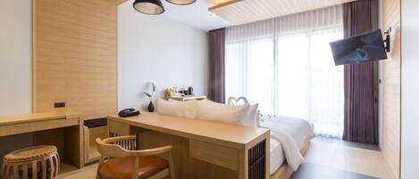 A modern hotel room with a wooden desk, a comfortable chair, a bed with a heart-shaped pillow, and a flat-screen TV mounted on the wall.