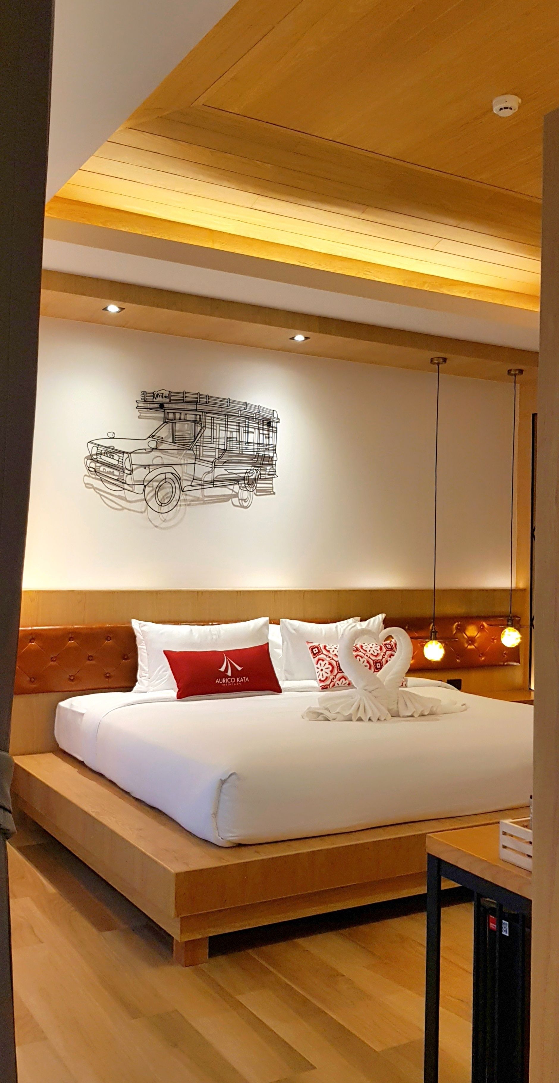 A modern hotel room with a wooden headboard, a bed with white bedding and red pillows, a wall-mounted artwork of a truck, and pendant lighting.