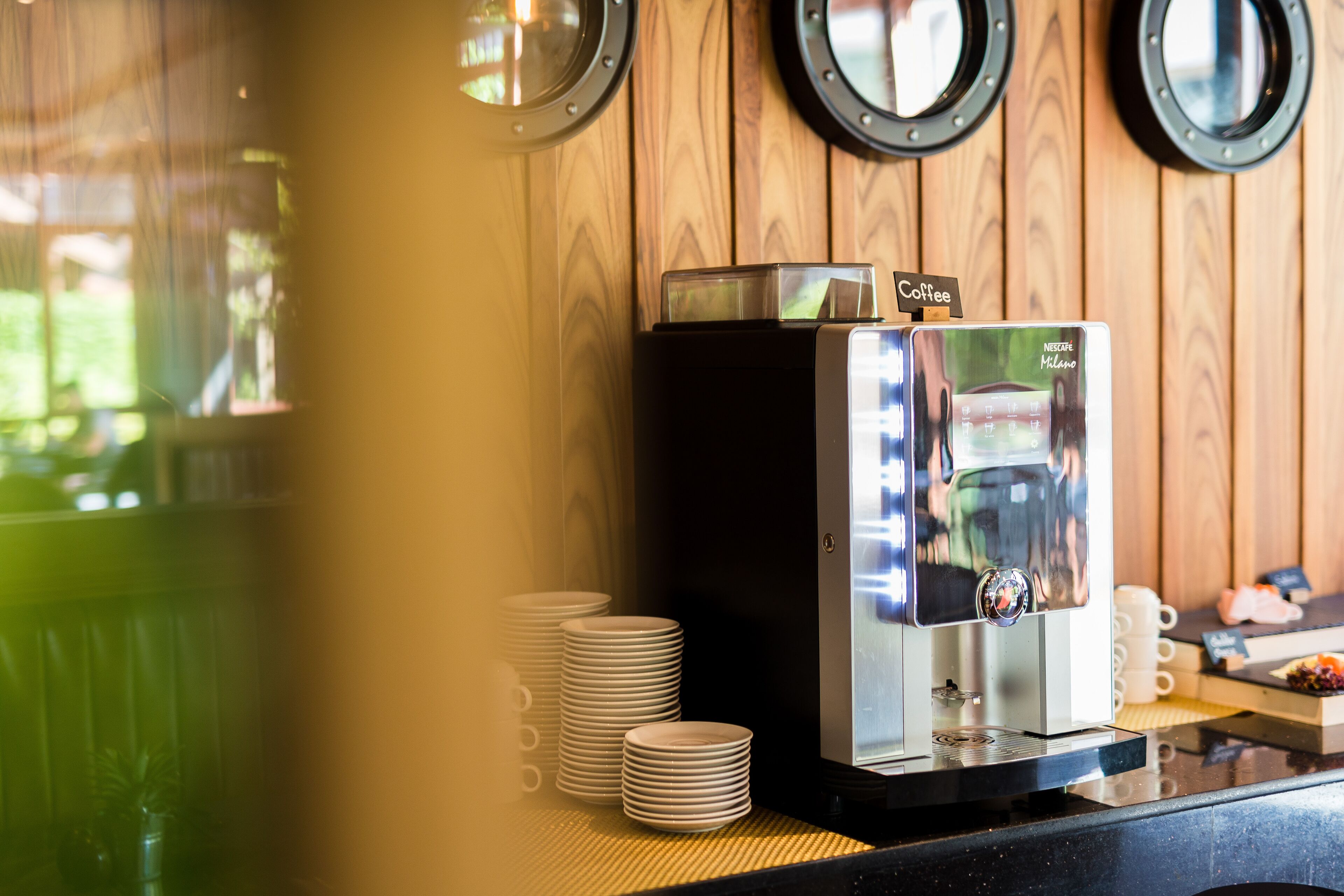 A coffee machine with a digital display, stacked white plates, and a wooden wall with circular windows.
