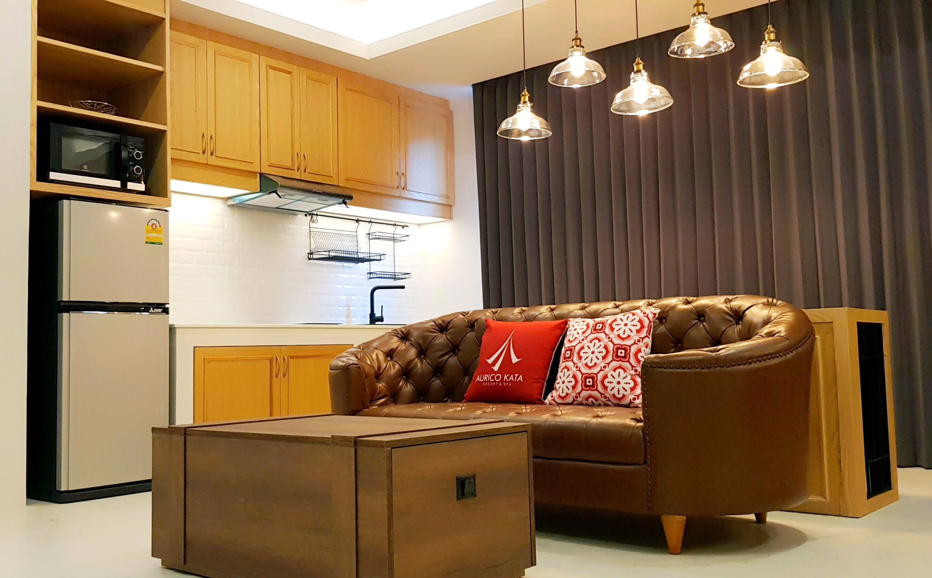 A modern living room with a leather sofa, a wooden coffee table, and a kitchen area with wooden cabinets and a refrigerator.