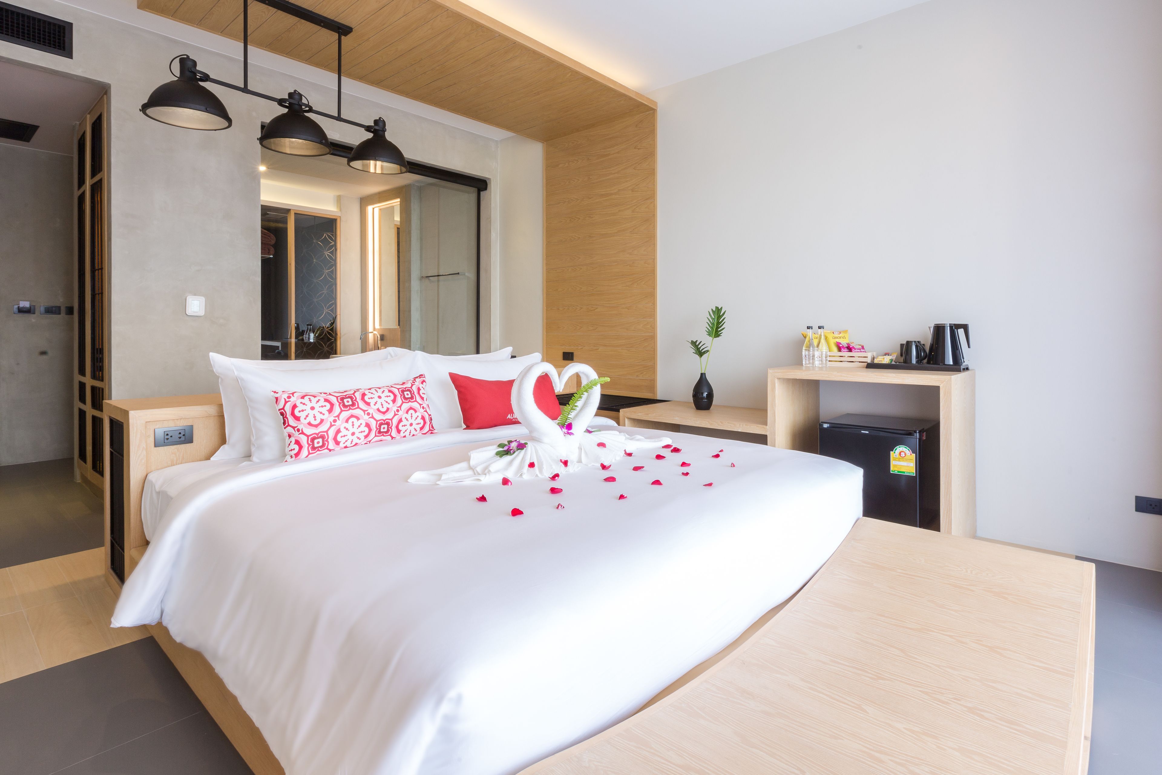 Ein modernes Hotelzimmer mit einem großen Bett, einem hölzernen Kopfteil, einem Nachttisch, einem Schreibtisch mit Stuhl, einem kleinen Kühlschrank und einem Spiegel.