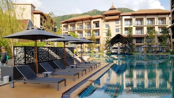 A resort pool area with lounge chairs and umbrellas, surrounded by a stone wall and a building with multiple balconies.