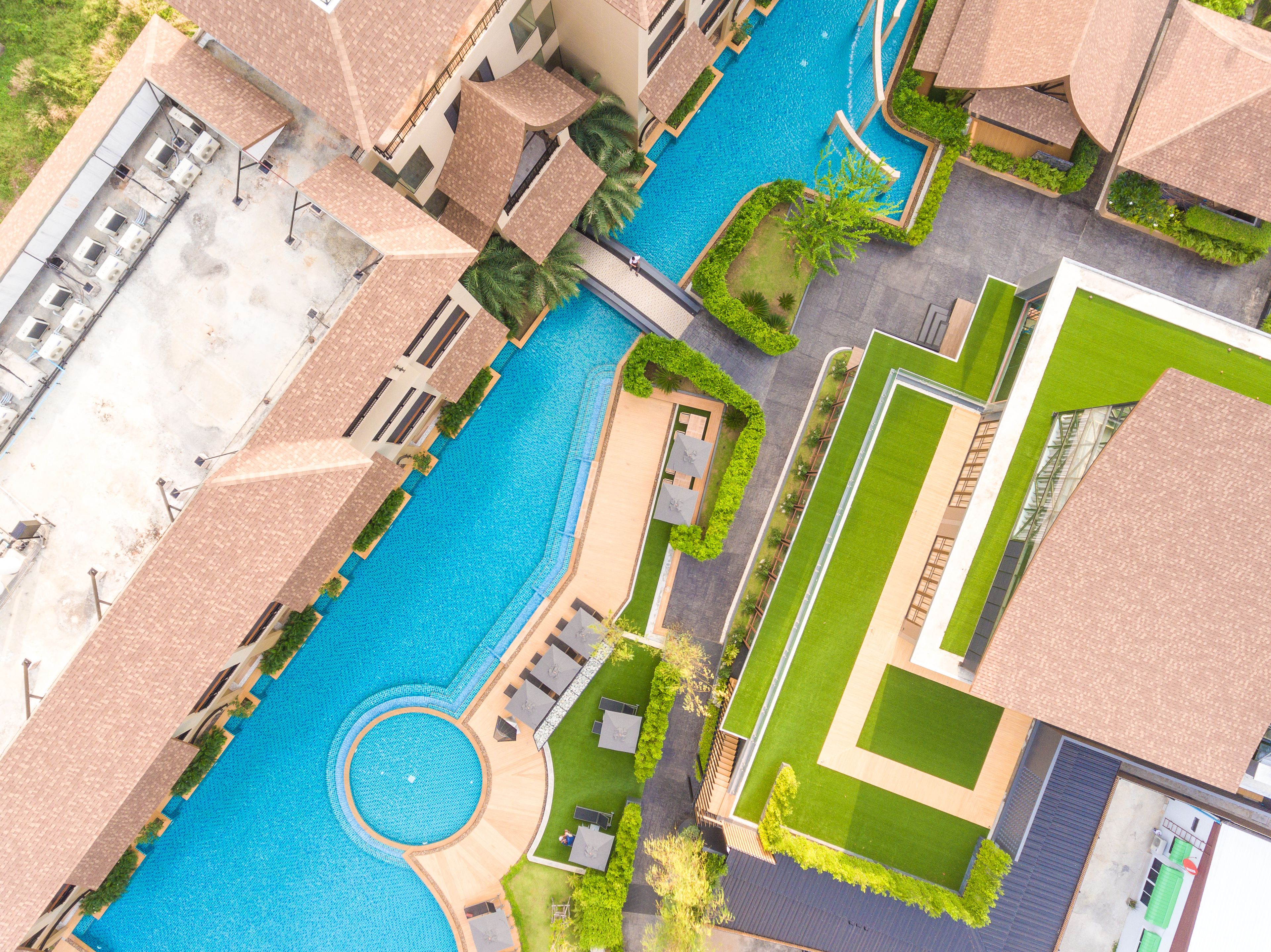 An aerial view of a resort with a central swimming pool, multiple buildings with tiled roofs, and well-maintained green lawns.