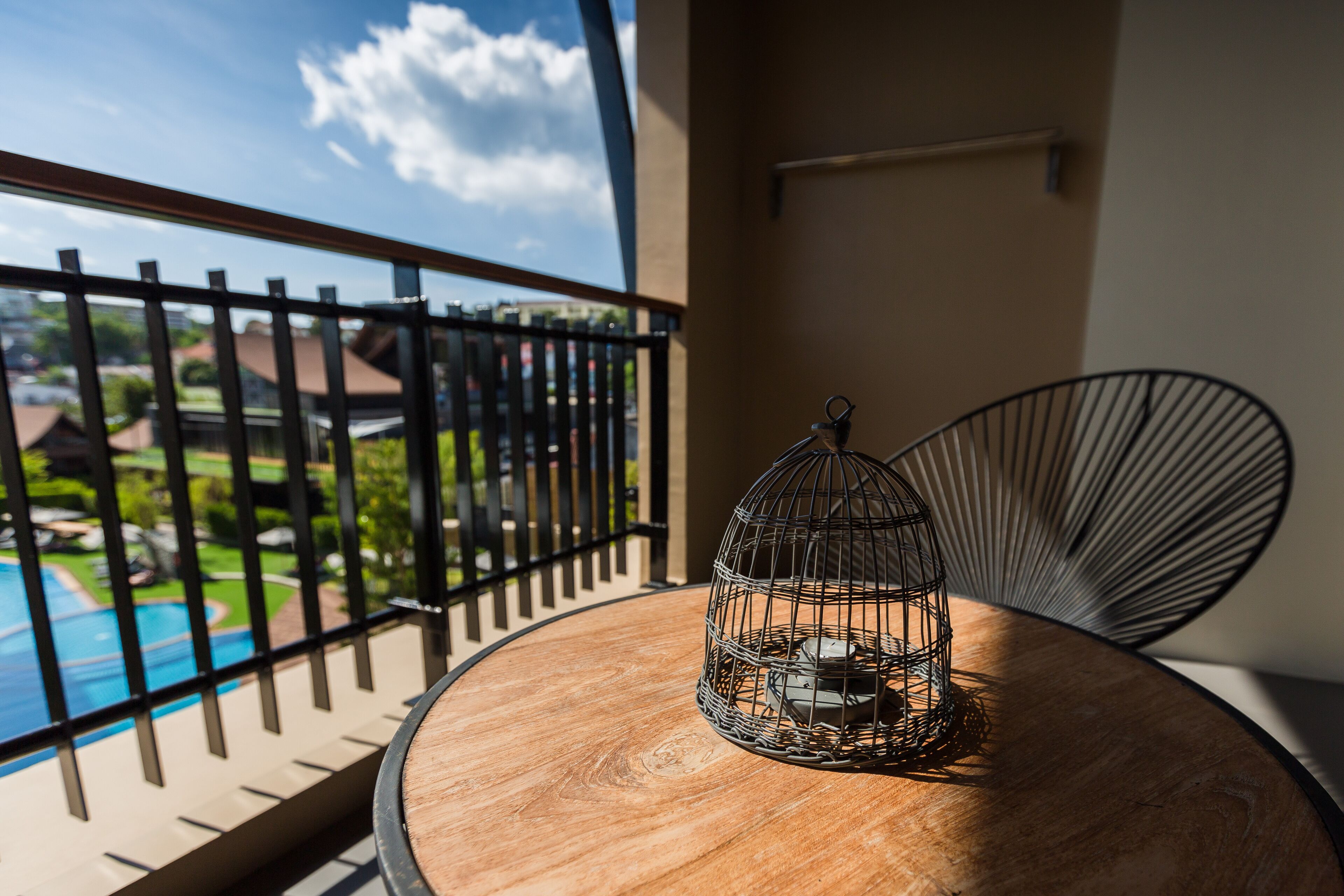 A round wooden table with a metal birdcage on it, a black metal chair, and a view of a swimming pool and greenery through a balcony railing.