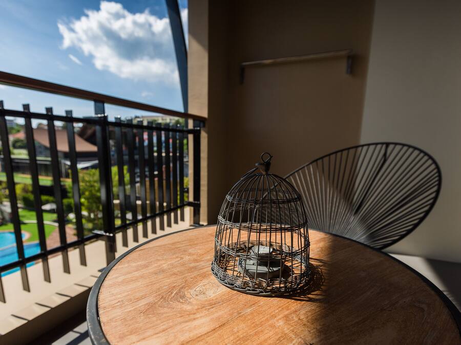 A round wooden table with a metal birdcage on it, a black metal chair, and a view of a swimming pool and greenery through a balcony railing.