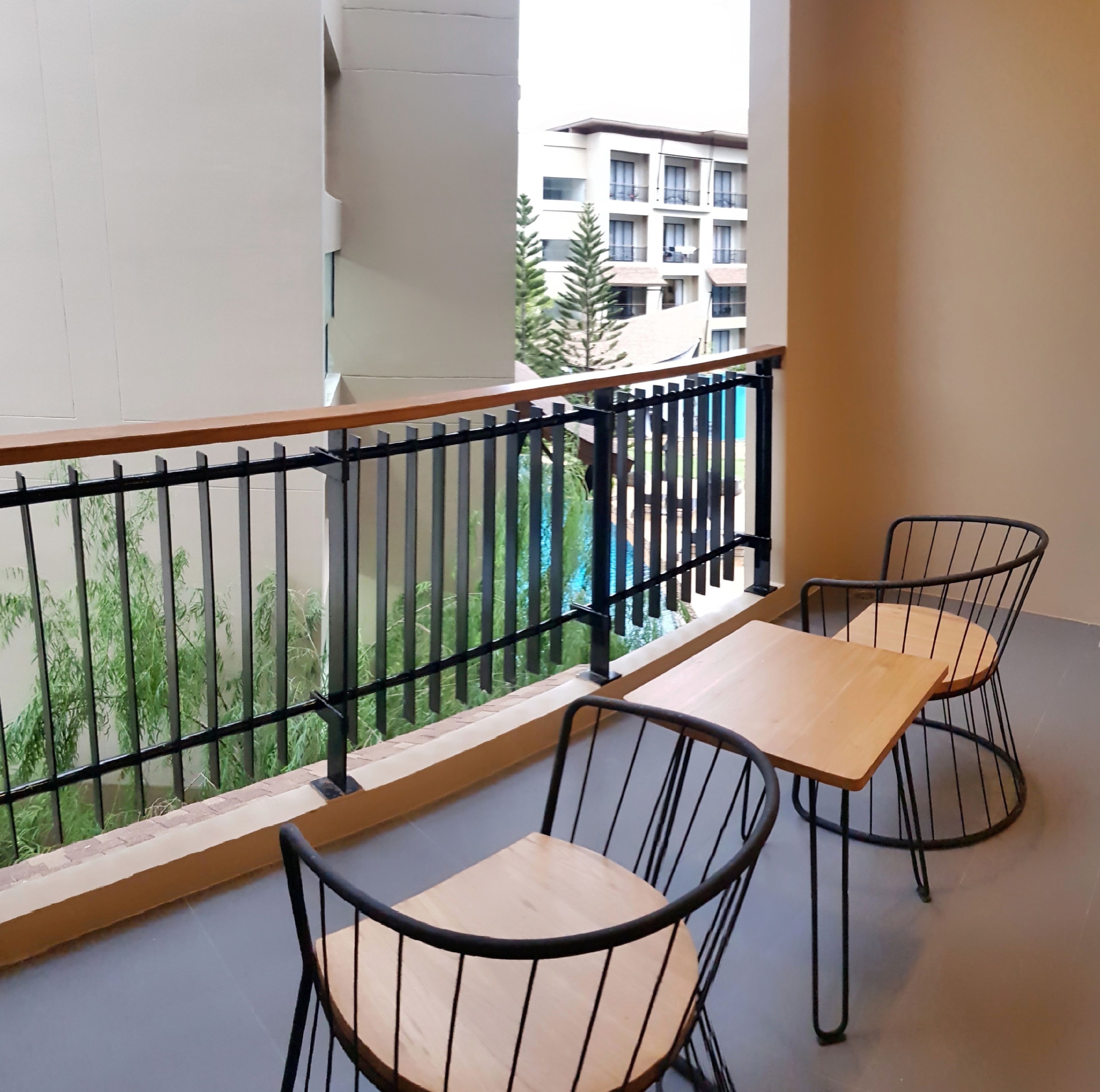 A balcony with a wooden table and two chairs, a metal railing, and a view of a building and greenery.