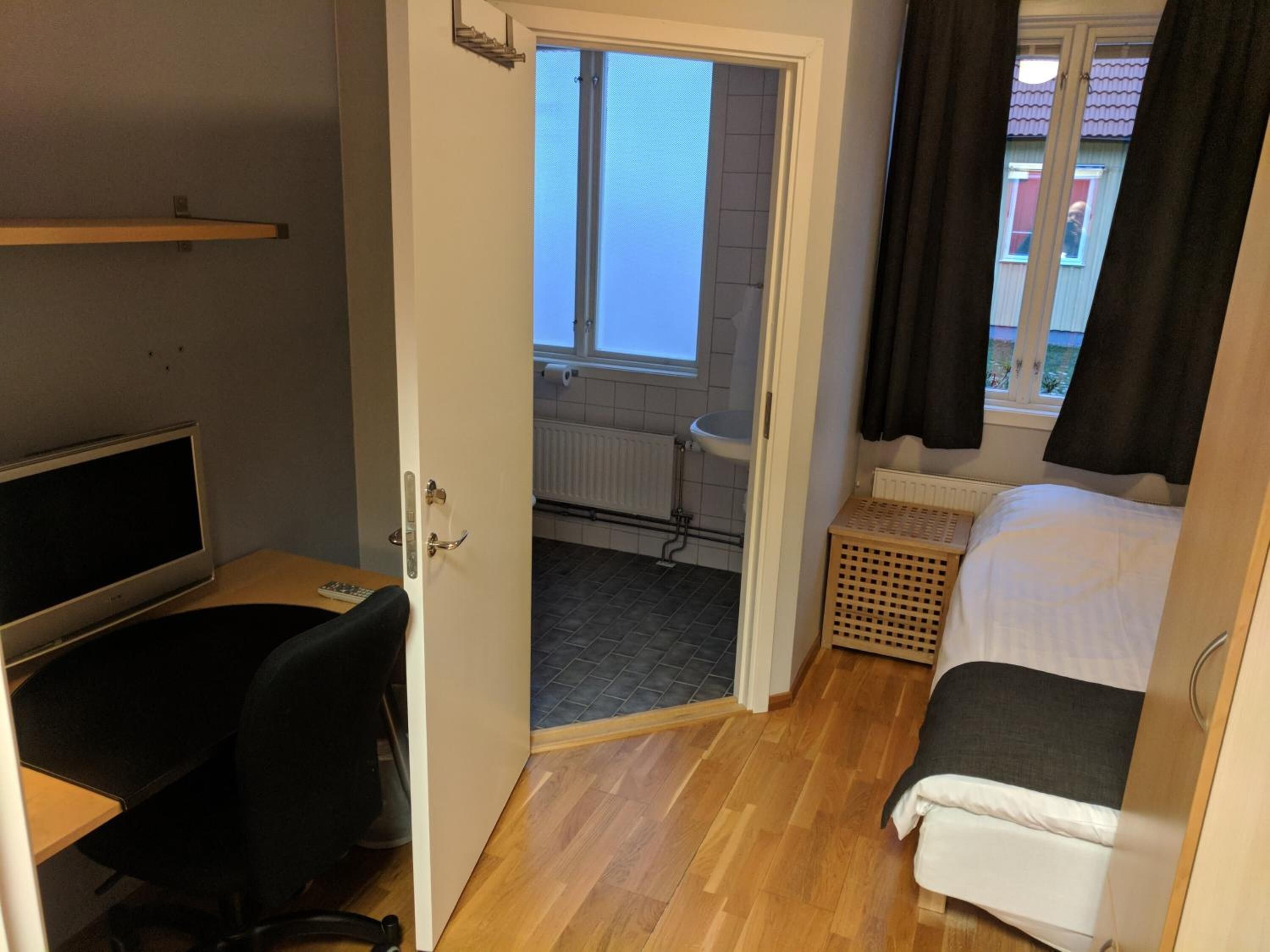 Habitació petita amb un llit, escriptori i cadira, televisió i finestra amb cortines.